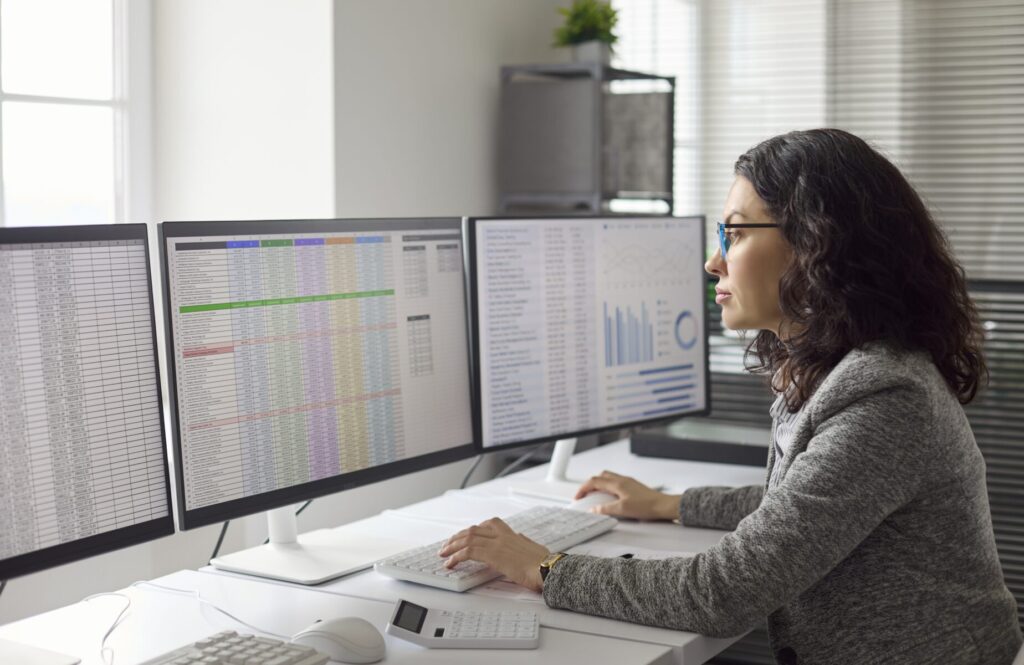 Profissional analisa planilhas e gráficos exibidos em três monitores em uma mesa de escritório.
